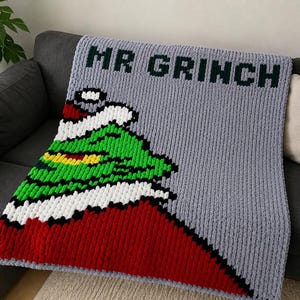 May include: A gray crocheted blanket featuring the text MR GRINCH in black letters at the top. The blanket displays a graphic of a green character wearing a red hat with white trim and a yellow collar, set against the textured gray background.