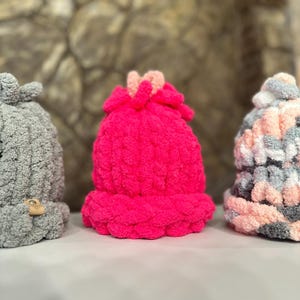 Op de afbeelding: Drie gebreide mutsen in verschillende kleuren en patronen. De mutsen zijn grijs, roze en een mix van grijs, roze en wit.