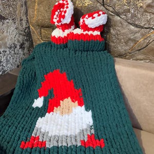 Può includere: Una coperta lavorata a mano di colore verde con un disegno di gnomo in rosso, bianco, grigio e beige. Due scarpine abbinate in maglia rossa e bianca sono sopra. La coperta e le scarpine sono realizzate in filato grosso.