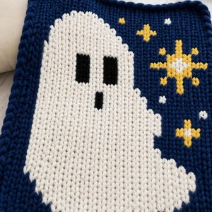 May include: A chunky knit blanket featuring a large white ghost centred on a deep navy blue background. The design includes yellow and white star motifs scattered to the right of the ghost. The blanket has a thick, textured yarn construction.