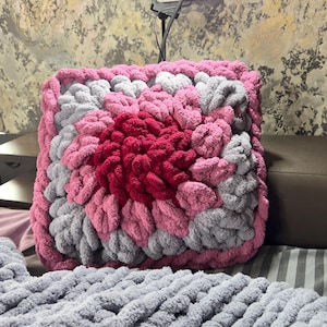Könnte beinhalten: Ein rosa und graues gestricktes Kissen mit einem großen, roten, blumenförmigen Design in der Mitte.
