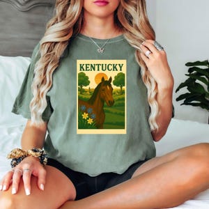 May include: Sage green t-shirt featuring a rectangular graphic with a brown horse, the word "Kentucky", a sunset, and a rural landscape. The graphic includes trees, fields, and flowers. The shirt is a solid color.