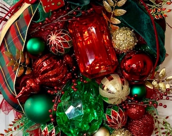 May include: A Christmas wreath decorated with red, green, and gold ornaments, including a large red jewel-shaped ornament and a green jewel-shaped ornament. The wreath is adorned with plaid ribbon, green velvet ribbon, and gold leaf sprigs.