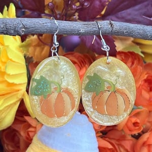 Puede incluir: Pendientes ovalados con diseño de calabaza. Los pendientes tienen un fondo amarillo brillante con una calabaza naranja y hojas verdes. Cuelgan de ganchos plateados. Un accesorio de temporada.