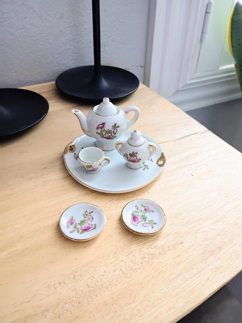 Pu&ograve; includere: Un servizio da t&egrave; in miniatura in porcellana bianca con disegni floreali rosa e bordi dorati. Il set comprende una teiera, zuccheriera, tazza e due piattini, disposti su un vassoio bianco. Esposto su una superficie di legno chiaro.