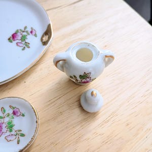 Pu&ograve; includere: Un servizio da t&egrave; in miniatura in porcellana con motivi floreali. Il set include una zuccheriera con coperchio e due piattini. I bordi dei piattini e della zuccheriera sono rifiniti con oro. Il motivo floreale presenta fiori rosa e verdi.