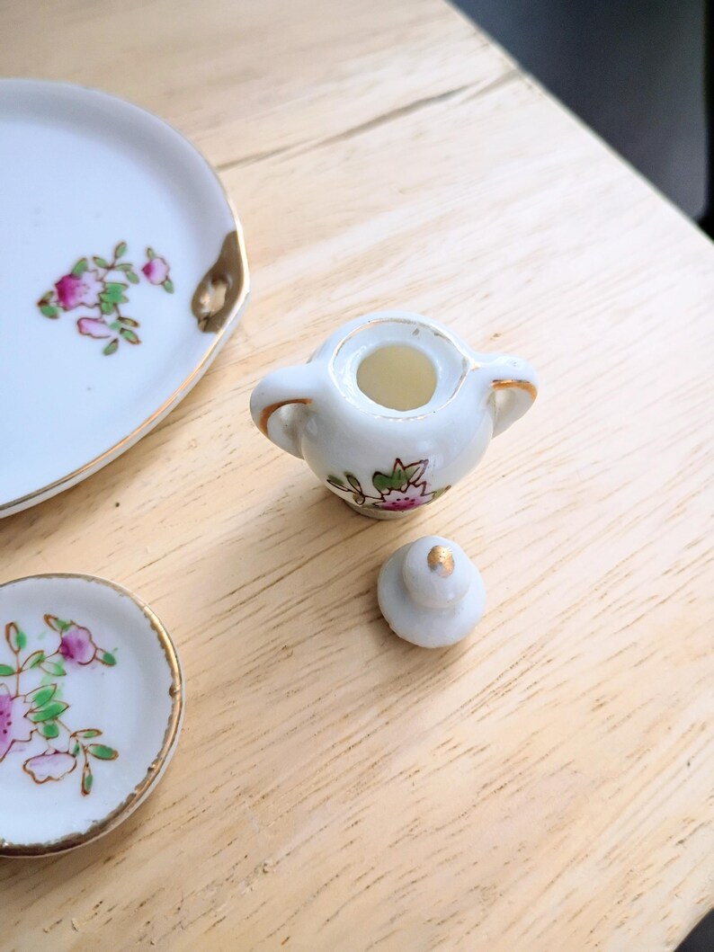 Pu&ograve; includere: Un servizio da t&egrave; in miniatura in porcellana con motivi floreali. Il set include una zuccheriera con coperchio e due piattini. I bordi dei piattini e della zuccheriera sono rifiniti con oro. Il motivo floreale presenta fiori rosa e verdi.