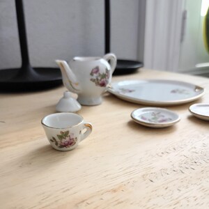 Pu&ograve; includere: Un servizio da t&egrave; in miniatura in porcellana con motivi floreali. Il set comprende una teiera, una tazza, un piattino e un piatto da portata. I pezzi sono bianchi con bordi dorati e motivi floreali rosa e verdi.