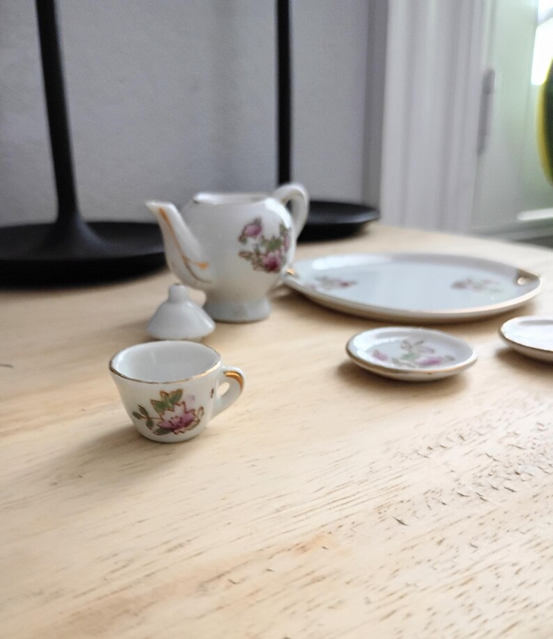 Pu&ograve; includere: Un servizio da t&egrave; in miniatura in porcellana con motivi floreali. Il set comprende una teiera, una tazza, un piattino e un piatto da portata. I pezzi sono bianchi con bordi dorati e motivi floreali rosa e verdi.