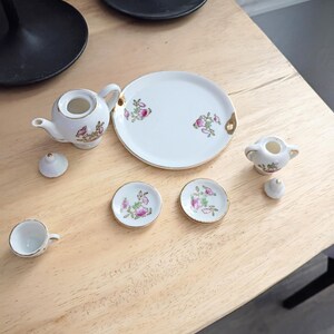 Pu&ograve; includere: Un servizio da t&egrave; in miniatura in porcellana bianca con disegni floreali e bordi dorati. Il set comprende una teiera, zuccheriera, piatto da portata, due piattini e una tazza da t&egrave;. Il servizio da t&egrave; &egrave; disposto su una superficie di legno chiaro.