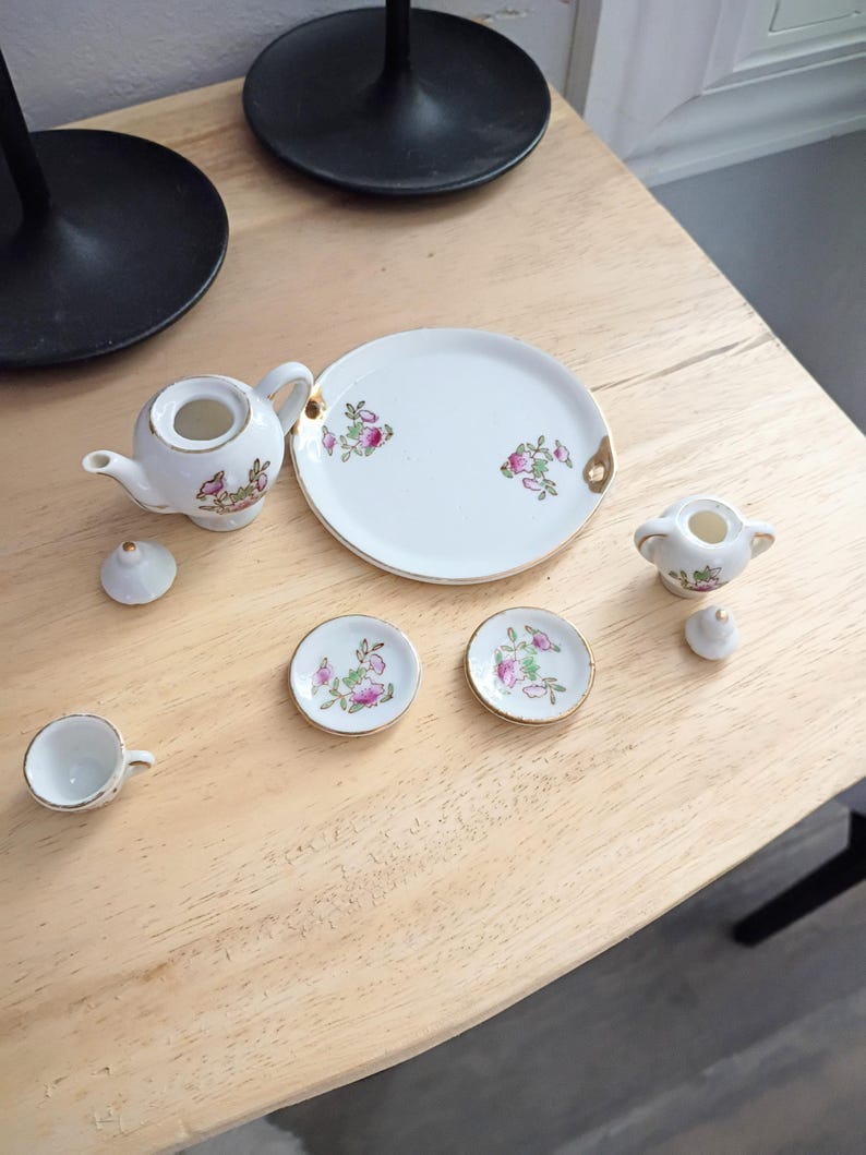 Pu&ograve; includere: Un servizio da t&egrave; in miniatura in porcellana bianca con disegni floreali e bordi dorati. Il set comprende una teiera, zuccheriera, piatto da portata, due piattini e una tazza da t&egrave;. Il servizio da t&egrave; &egrave; disposto su una superficie di legno chiaro.