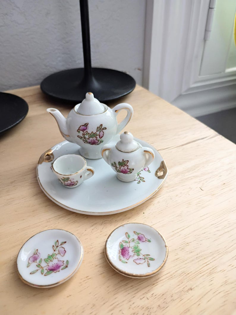 Pu&ograve; includere: Un servizio da t&egrave; in miniatura in porcellana bianca con disegni floreali rosa e bordi dorati. Il set comprende una teiera, zuccheriera, tazza da t&egrave; e due piattini, il tutto disposto su un vassoio rotondo.