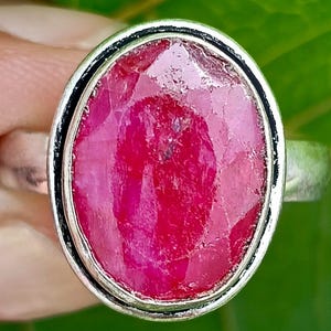 Könnte beinhalten: Ein Silberring mit einem großen, oval geschliffenen Rubin. Der Rubin ist ein leuchtendes Rot mit sichtbaren Facetten, gefasst in einer Silberfassung. Der Ring hat ein schlichtes, poliertes Silberband.
