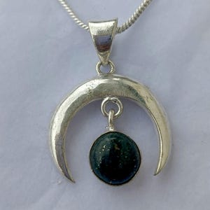 May include: A silver necklace with a crescent moon pendant and a dark green spherical stone. The pendant hangs from a fine silver chain. The pendant has a polished, metallic finish. The stone is a dark green colour.