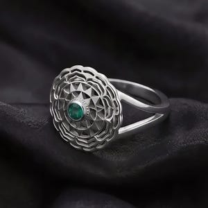 May include: A silver-toned ring featuring a circular, multi-layered geometric face with a scalloped outer edge. The center of the ring is set with a small, round, deep green gemstone. The band is split into two thin, polished silver-toned arms.
