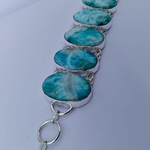 May include: A silver bracelet featuring oval-shaped, light blue gemstones with white marbling. The gemstones are connected by silver chain links. The bracelet is displayed on a white surface.
