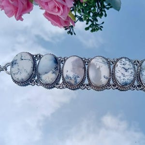 May include: Silver-toned bracelet featuring six oval-shaped, white gemstones with black and gray dendritic patterns. The bracelet has a toggle clasp and is displayed on a reflective surface with pink flowers in the background.