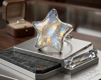 Puede incluir: Un anillo de plata con una gran piedra preciosa iridiscente en forma de estrella con tonos azules y dorados. El anillo se exhibe sobre un soporte escalonado de metal y piedra con la inscripción ASTRAA JEWELERS, junto a un pequeño joyero de madera.