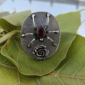 May include: Oval-shaped silver ring featuring a spider design with a central red gemstone and a rose detail. The ring has a textured surface and is set against a green leaf.
