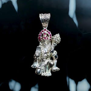 May include: A silver pendant depicting a deity, adorned with red gemstones on the crown. The pendant features intricate details and is suspended from a textured bail. The figure is in a kneeling position, holding an object.