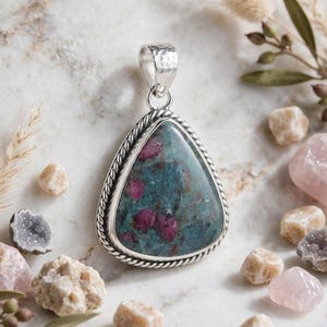 May include: A triangular pendant featuring a polished ruby in zoisite gemstone with deep red and teal green inclusions. The stone is set in a silver frame with a twisted rope border and a hammered silver bail at the top for a necklace chain.