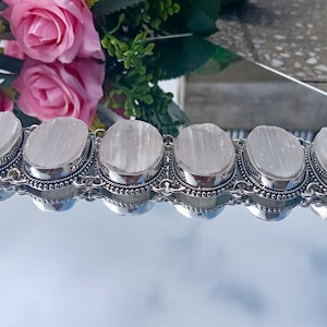 May include: A silver bracelet featuring six oval, light-colored stones. Each stone is set in a silver bezel with a beaded border. The bracelet is displayed against a reflective surface, with pink roses in the background.