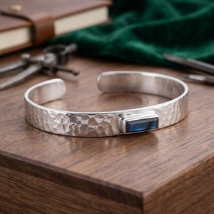 May include: A silver cuff bracelet featuring a hammered texture and a single rectangular blue labradorite gemstone set in a smooth metal bezel. The open-ended band has a polished finish and displays a distinct iridescent sheen on the stone.