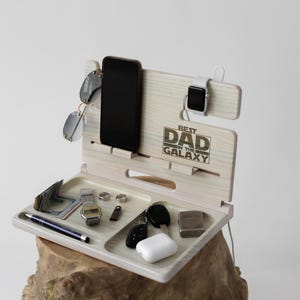 May include: Wooden phone stand with a tray for holding keys, a watch, and other small items. The stand has a slot for a phone and a watch holder. The stand is engraved with the text "Best Dad in the Galaxy".