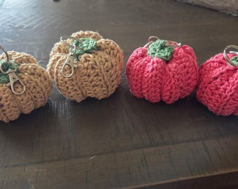 May include: Four handmade crochet pumpkin keychains in shades of tan and red. Each pumpkin has a green leaf and a metal clasp or ring. The keychains are arranged on a dark wooden surface.