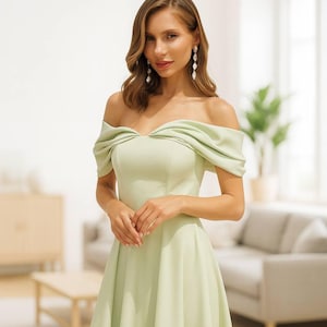 Może przedstawiać: Jasnozielona sukienka z odkrytymi ramionami. Sukienka ma dopasowany gorset i zwiewną spódnicę. Modelka ma na sobie długie, wiszące kolczyki. Tło obejmuje sofę, stolik kawowy i roślinę domową.