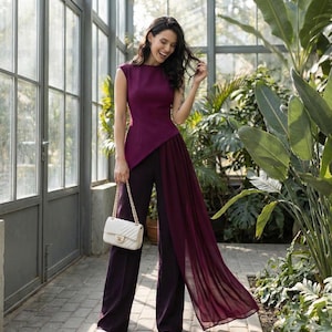 May include: A plum-coloured sleeveless top with an asymmetrical design and a flowing chiffon overlay, paired with wide-leg trousers. A white quilted handbag with a gold chain strap completes the ensemble. The outfit is suitable for formal events.