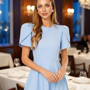 May include: A light blue dress with short sleeves and a fitted bodice. The dress has a flared skirt and is worn in a restaurant setting. The model is wearing earrings and has long, wavy blonde hair.