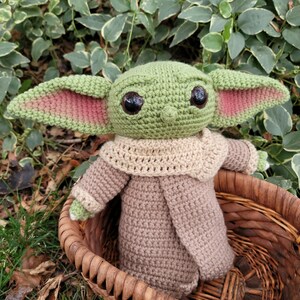 Może przedstawiać: Pluszowa zabawka Baby Yoda wykonana na szydełku w kolorze zielonym i brązowym, ubrana w brązowy płaszcz, siedząca w wiklinowym koszu. Zabawka ma duże brązowe oczy i r&oacute;żowe uszy.