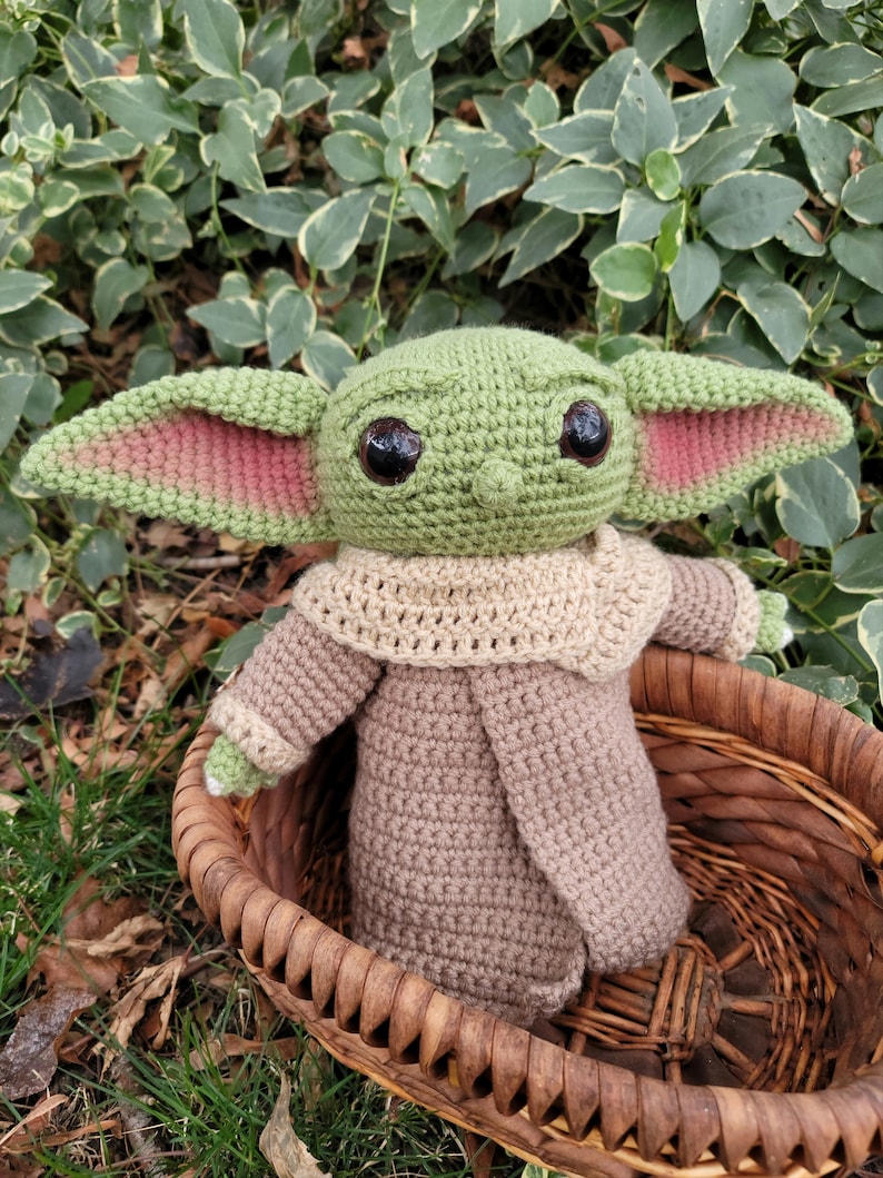 Może przedstawiać: Pluszowa zabawka Baby Yoda wykonana na szydełku w kolorze zielonym i brązowym, ubrana w brązowy płaszcz, siedząca w wiklinowym koszu. Zabawka ma duże brązowe oczy i r&oacute;żowe uszy.