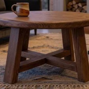 May include: A round wooden coffee table with a dark brown finish, featuring four thick, angled legs connected by a central cross-brace support structure. A two-toned ceramic mug with a tan top and brown base sits on the table surface.