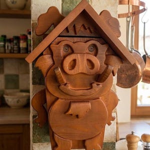 Könnte beinhalten: Eine wandmontierte Holzschnitzerei in Form eines stilisierten Warzenschweins mit einer Dachkonstruktion über dem Kopf. Das braun gebeizte Holz zeigt geschnitzte Gesichtsdetails wie Rüssel, Stoßzähne und Ohren sowie einen runden Körper.