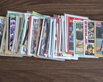 May include: A collection of baseball trading cards, many in protective sleeves, featuring various players and teams. The cards are arranged in a slightly fanned-out pile on a wooden surface. Some cards are visible, including one with "Nolan Ryan" printed on it.