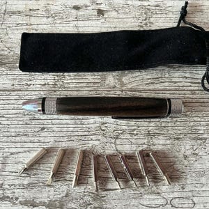 May include: A dark wood pen with silver detailing and a set of interchangeable tips. The pen is presented with a black velvet pouch. The pen and tips are arranged on a light wood-grain surface.