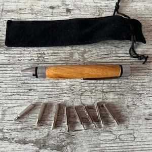 May include: A wooden pen with silver detailing and a set of small silver tools, alongside a black drawstring pouch, are arranged on a light wood-grain surface. The pen features a natural wood finish and a silver tip.