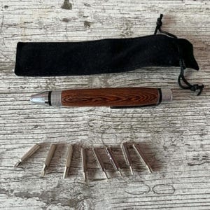 May include: A wooden pen with a silver tip and accents, a black velvet pouch, and several silver pen tips. The pen features a dark brown wood grain pattern.