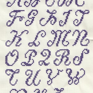 Könnte beinhalten: Ein violett besticktes Alphabet mit kursivem Schriftzug auf einem weißen Stoffhintergrund.