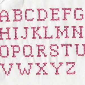 May include: A pink embroidered alphabet on a white fabric background. The letters are stitched in a simple cross-stitch pattern.