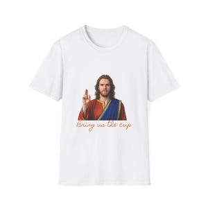 May include: White t-shirt featuring a graphic of a man in a red and blue robe, raising his hand. The text "Bring us the cup" is written in a cursive font below the image. The shirt is a casual wear item.