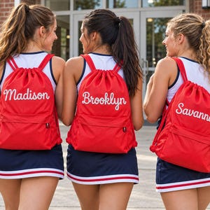 May include: Three bright red drawstring backpacks made of mesh fabric, each featuring a white front zipper pocket and a personalized name embroidered in white script: Madison, Brooklyn, and Savannah, worn over white and navy athletic uniforms.
