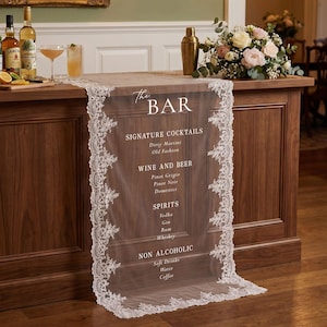 May include: A wooden bar with a sheer overlay featuring the text "The BAR" and a list of drinks. The overlay has a delicate lace trim. Bottles of liquor, cocktail glasses, and a cocktail shaker are on the bar. A floral arrangement is also present.