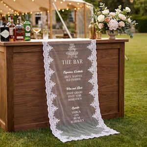 May include: A wooden bar with a white lace runner featuring the text "THE BAR" and a list of drinks. Bottles of liquor and a cocktail shaker sit on top. A floral arrangement and string lights are in the background.