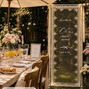 K&ouml;nnte beinhalten: Ein hohes, rechteckiges Willkommensschild aus cremefarbener Spitze f&uuml;r eine Brautparty mit der Aufschrift 'WELCOME TO Olivia's BRIDAL SHOWER' und 'JUNE 14, 2026', aufgeh&auml;ngt an einem Metallrahmen mit B&auml;ndern, Ma&szlig;e 75 x 200 cm.