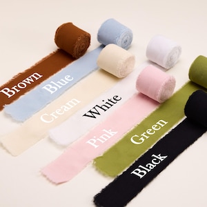 K&ouml;nnte beinhalten: Sieben Stoffbandrollen mit ausgefransten Kanten, die in einer diagonalen Linie angeordnet sind. Die Farben umfassen Braun, Blau, Creme, Wei&szlig;, Rosa, Gr&uuml;n und Schwarz. Jedes Band ist mit dem entsprechenden Farbnamen in wei&szlig;em oder schwarzem Text beschriftet.