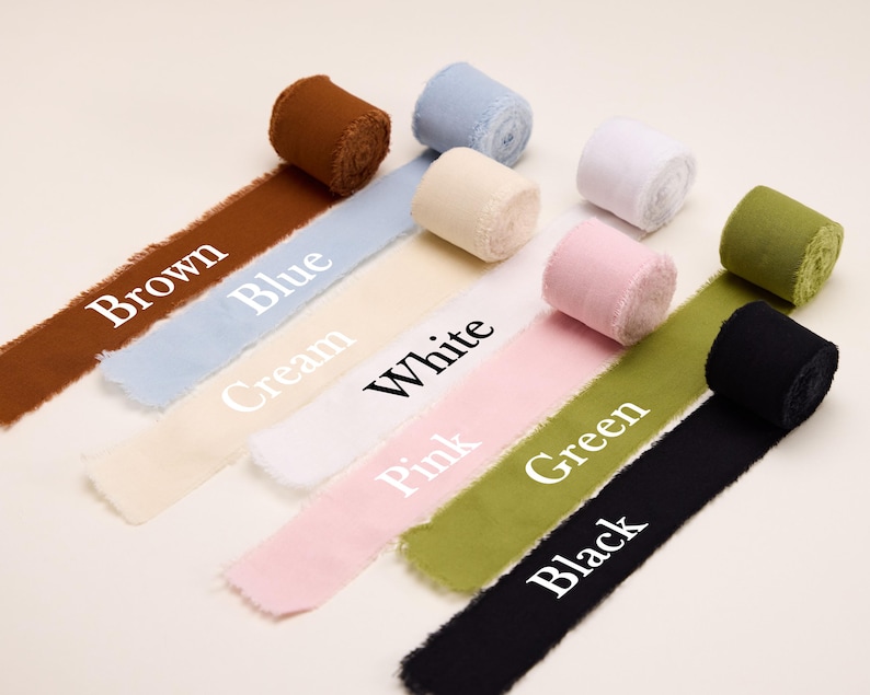 K&ouml;nnte beinhalten: Sieben Stoffbandrollen mit ausgefransten Kanten, die in einer diagonalen Linie angeordnet sind. Die Farben umfassen Braun, Blau, Creme, Wei&szlig;, Rosa, Gr&uuml;n und Schwarz. Jedes Band ist mit dem entsprechenden Farbnamen in wei&szlig;em oder schwarzem Text beschriftet.
