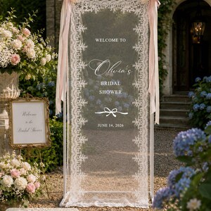 K&ouml;nnte beinhalten: Ein rechteckiger wei&szlig;er Metallrahmen mit einer halbtransparenten wei&szlig;en Spitzenbespannung, bedruckt mit WELCOME TO Olivia's BRIDAL SHOWER JUNE 14, 2026 und einer Schleifengrafik. Der Rahmen ist mit hellrosa B&auml;ndern verziert und misst 75 x 200 cm.